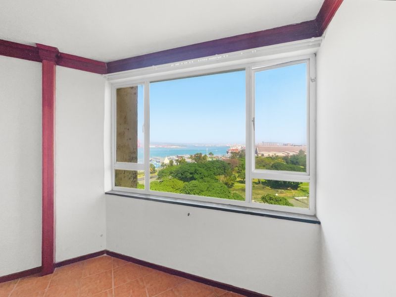An empty living area featuring terracotta floor tiles, decorative timber trim, and a large window offering expansive coastal and park views.
