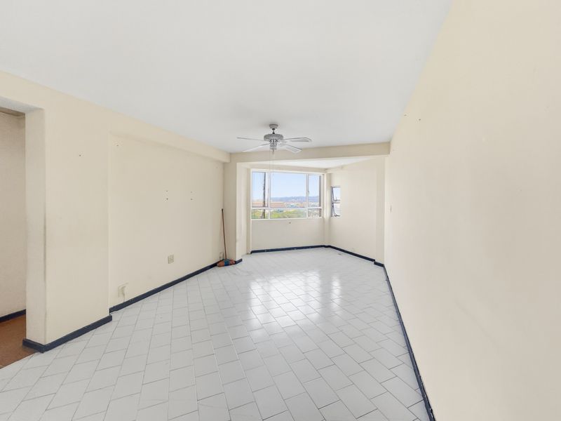 A spacious, empty living area featuring white tiled flooring, a ceiling fan, and large windows providing natural light and views.