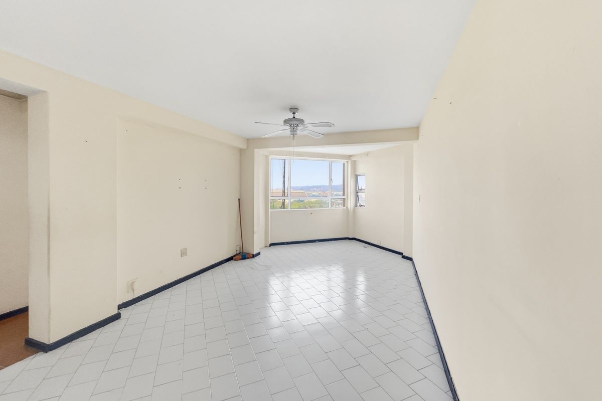 A spacious, empty living area featuring white tiled flooring, a ceiling fan, and large windows providing natural light and views.