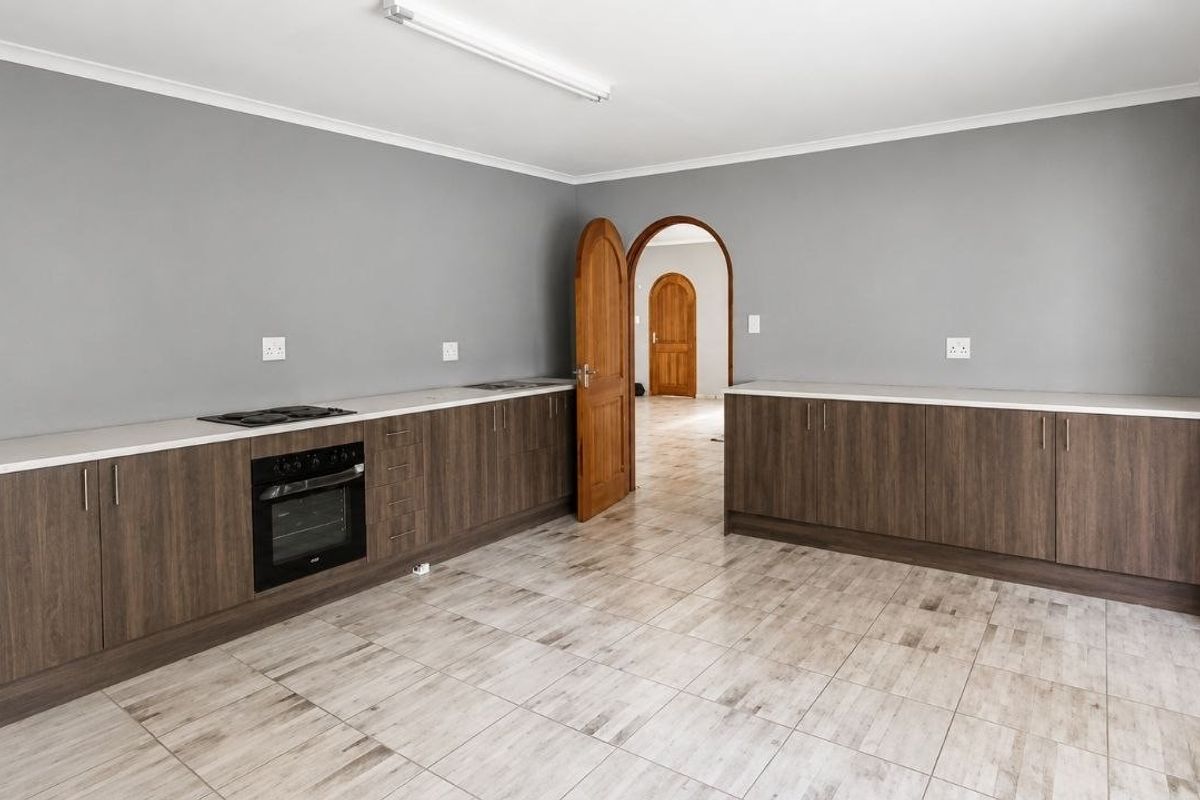 A clean, empty kitchen featuring modern wood-finish cabinetry, light countertops, integrated oven and hob, and neutral tiled flooring with an arched doorway.
