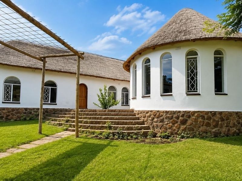A well-maintained rear exterior view featuring a unique thatched roof design, stone cladding, arched windows, and a timber pergola leading to stone steps.