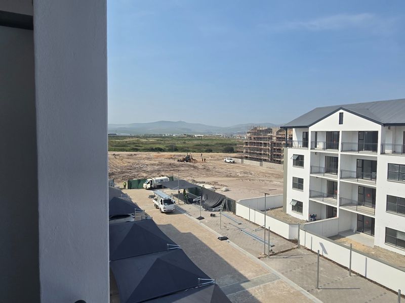 An elevated view from a balcony overlooking a modern apartment complex, paved parking area with carports, and surrounding development land.