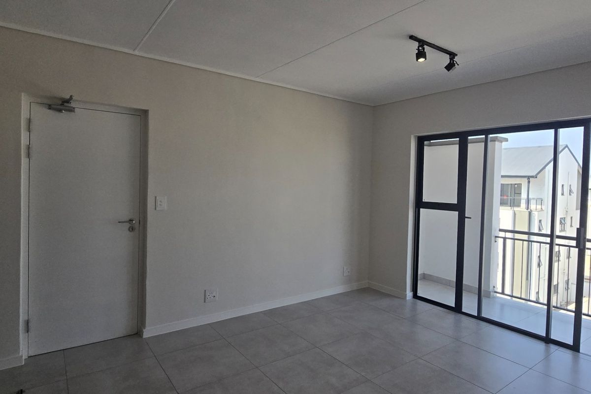 A bright, empty living area featuring neutral tiled flooring, modern track lighting, and a large sliding glass door leading to an outdoor balcony.
