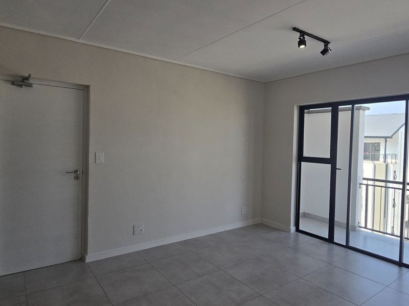 A bright, empty living area featuring neutral tiled flooring, modern track lighting, and a large sliding glass door leading to an outdoor balcony.