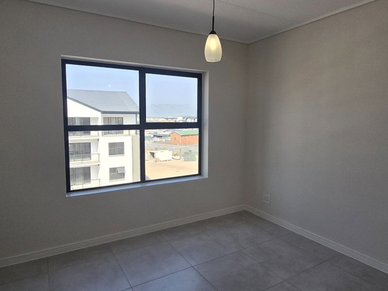 A bright, empty bedroom featuring neutral tiled flooring, modern aluminium window frames, and a simple pendant light fixture.