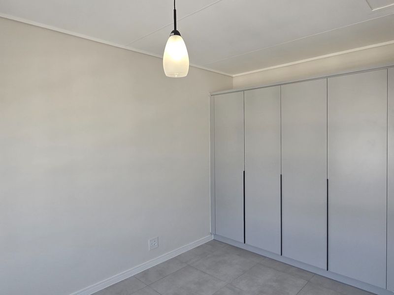 A bright, empty bedroom featuring modern built-in cupboards, neutral tiled flooring, and a contemporary pendant light fixture, with an adjacent kitchen view.