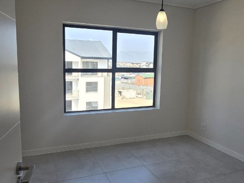 A bright, empty bedroom featuring modern grey floor tiles, a single pendant light, and a large window with views of the surrounding apartment complex.