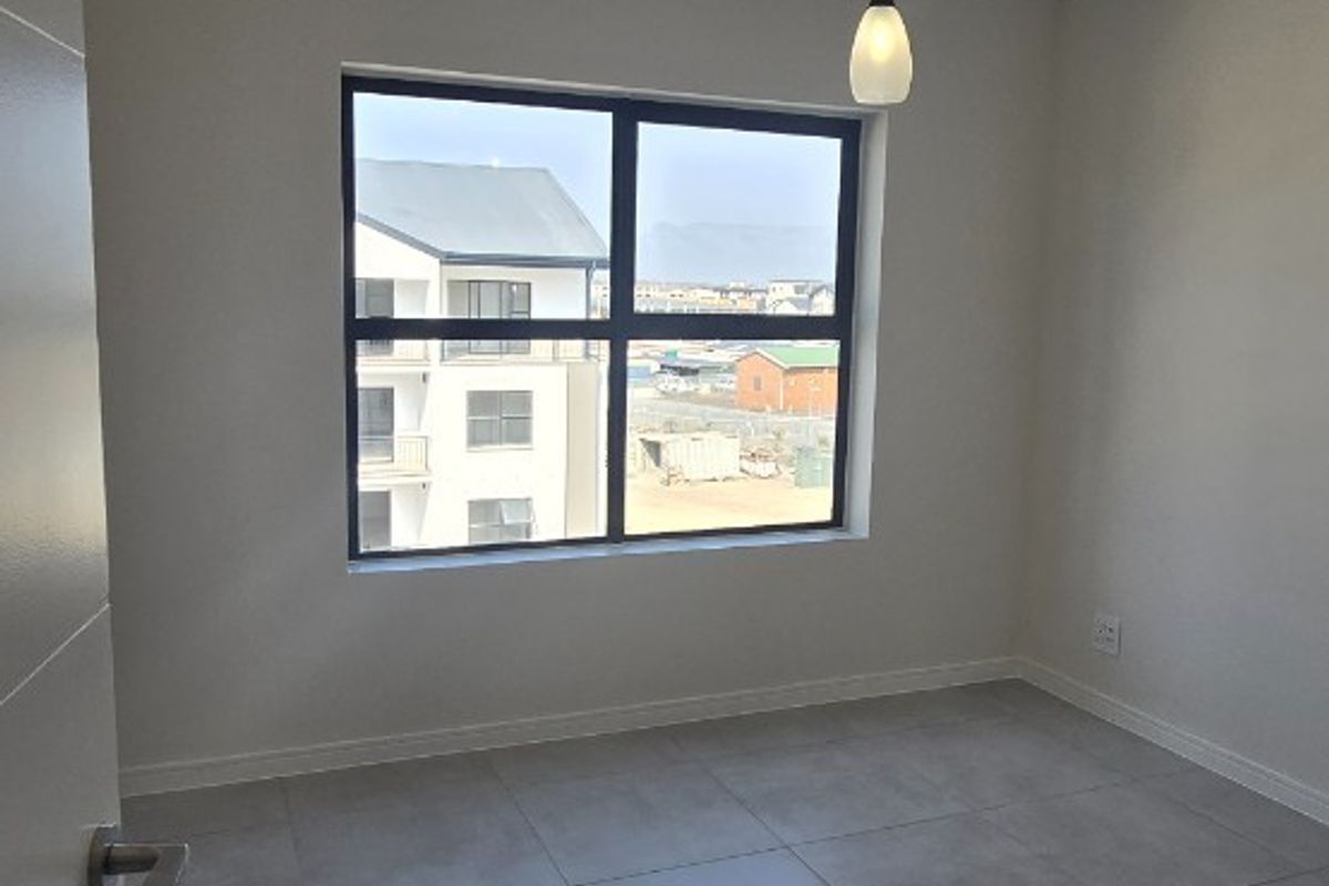 A bright, empty bedroom featuring modern grey floor tiles, a single pendant light, and a large window with views of the surrounding apartment complex.