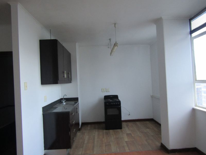 A compact, empty kitchen featuring dark cabinetry, a stainless steel sink, and a freestanding stove, with laminate flooring and natural light from a large window.
