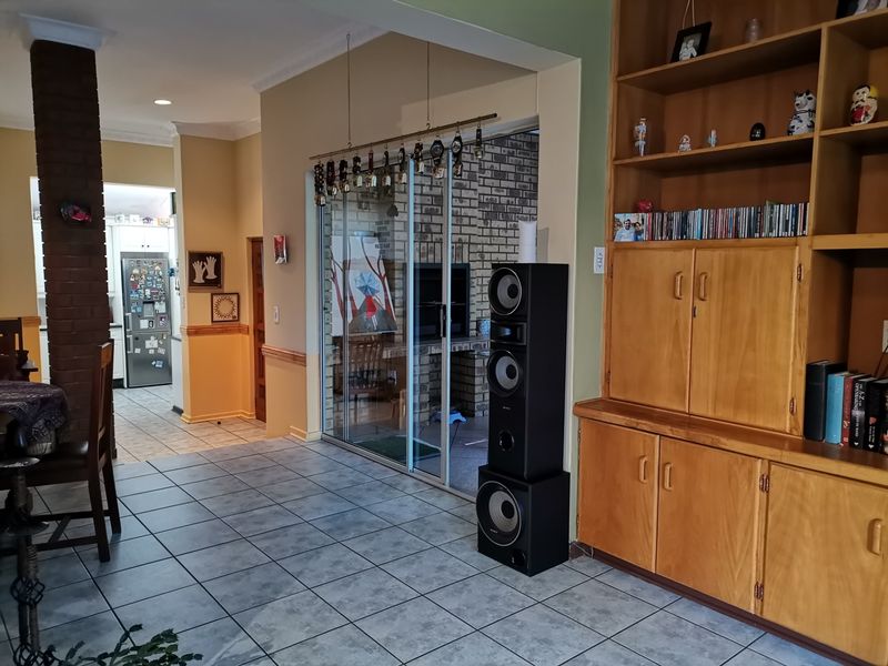 Open plan living area featuring tiled floors, a built-in wooden shelving unit, and a brick feature pillar, with a view into the kitchen area.