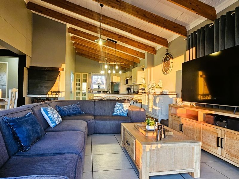 Spacious open-plan living area featuring exposed timber ceiling beams, tiled flooring, and a cozy built-in fireplace, seamlessly connecting to the kitchen.