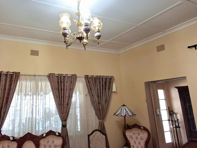 A traditional living room featuring a decorative chandelier, ceiling tiles, and coving, with access to an adjacent room through a doorway.