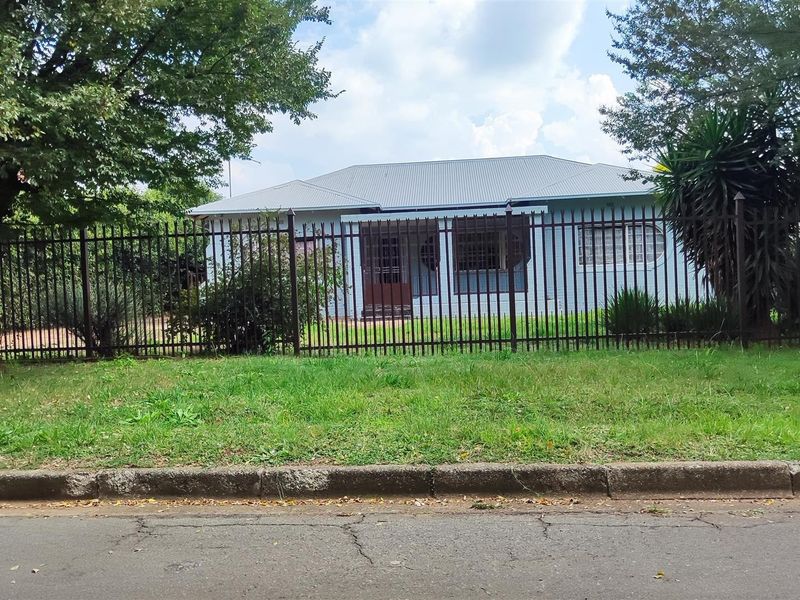 Front view of a single-story residential house featuring a metal roof and secure perimeter fencing, set back from the street.