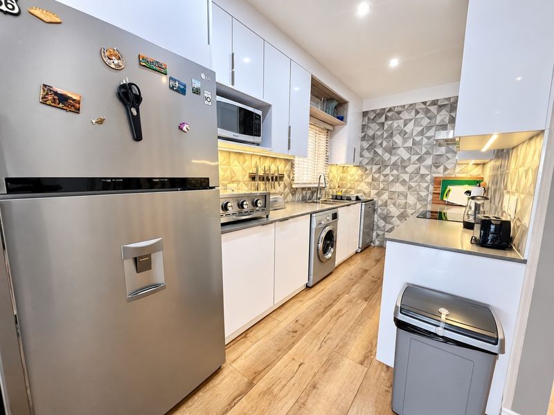 Modern kitchen featuring sleek white cabinetry, stainless steel countertops, integrated appliances, a geometric tiled backsplash, and warm wood-look laminate flooring.