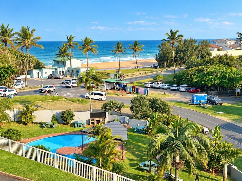 Elevated coastal view overlooking a swimming pool, landscaped garden, and the nearby beach and ocean.