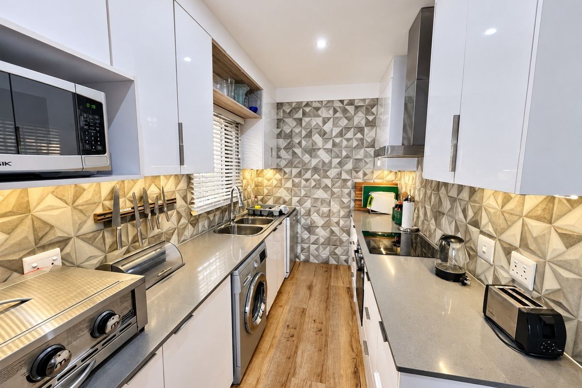 Modern galley kitchen featuring high-gloss white cabinetry, stone countertops, geometric tiled backsplash, and integrated appliances with warm under-cabinet lighting.