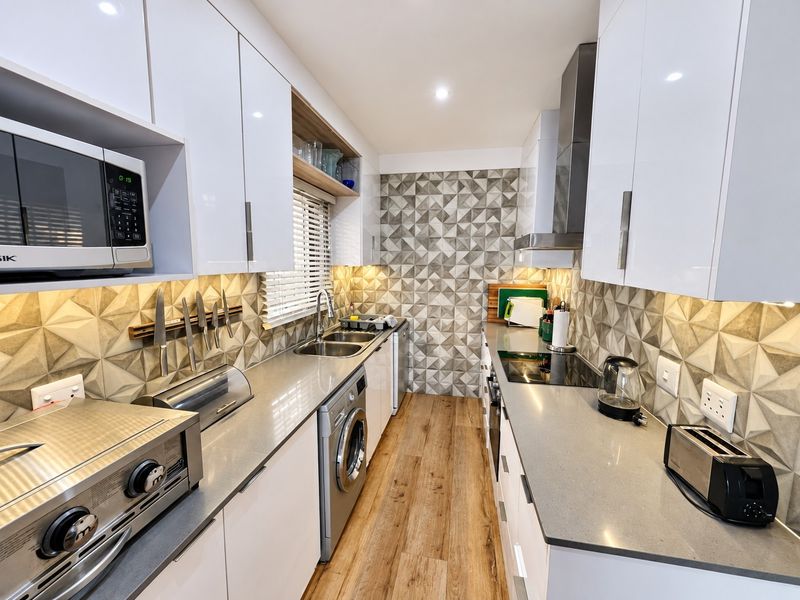 Modern galley kitchen featuring high-gloss white cabinetry, stone countertops, geometric tiled backsplash, and integrated appliances with warm under-cabinet lighting.