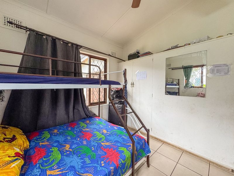 A bedroom featuring built-in cupboards, tiled flooring, and a ceiling fan. The room includes security bars on the windows and a wall-mounted mirror.
