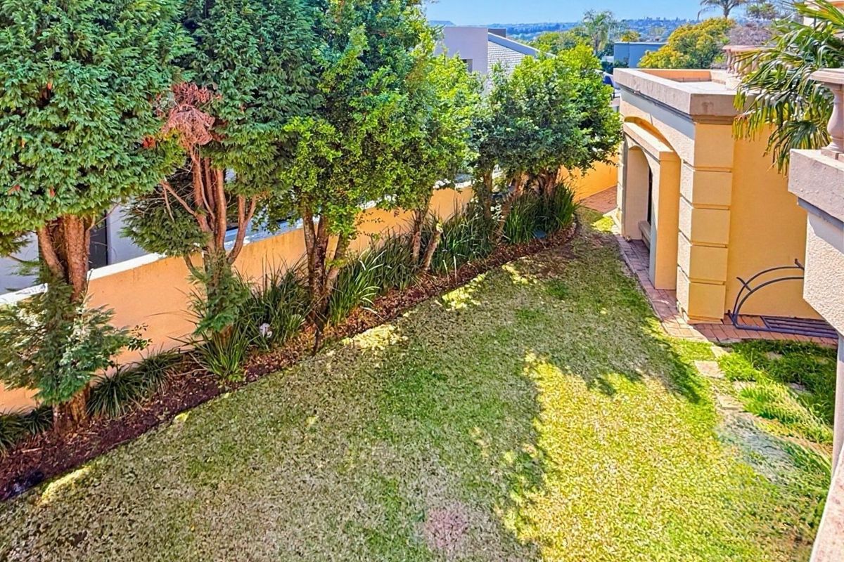 An elevated view of a private rear garden featuring a manicured lawn, mature perimeter trees, and a paved walkway adjacent to the building structure.