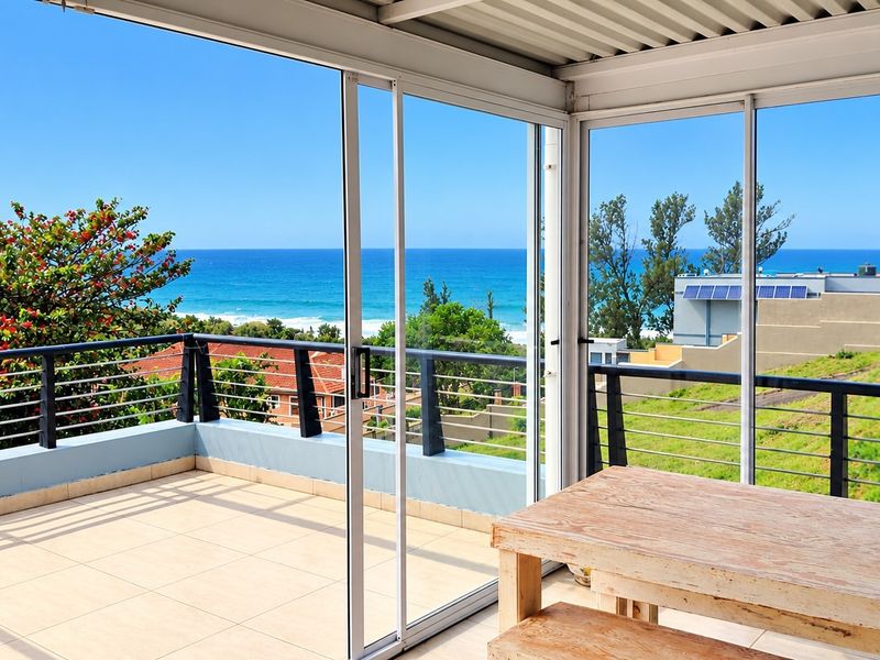 Spacious covered balcony featuring tiled flooring, modern sliding glass doors, and a metal balustrade, offering expansive ocean and coastal views.