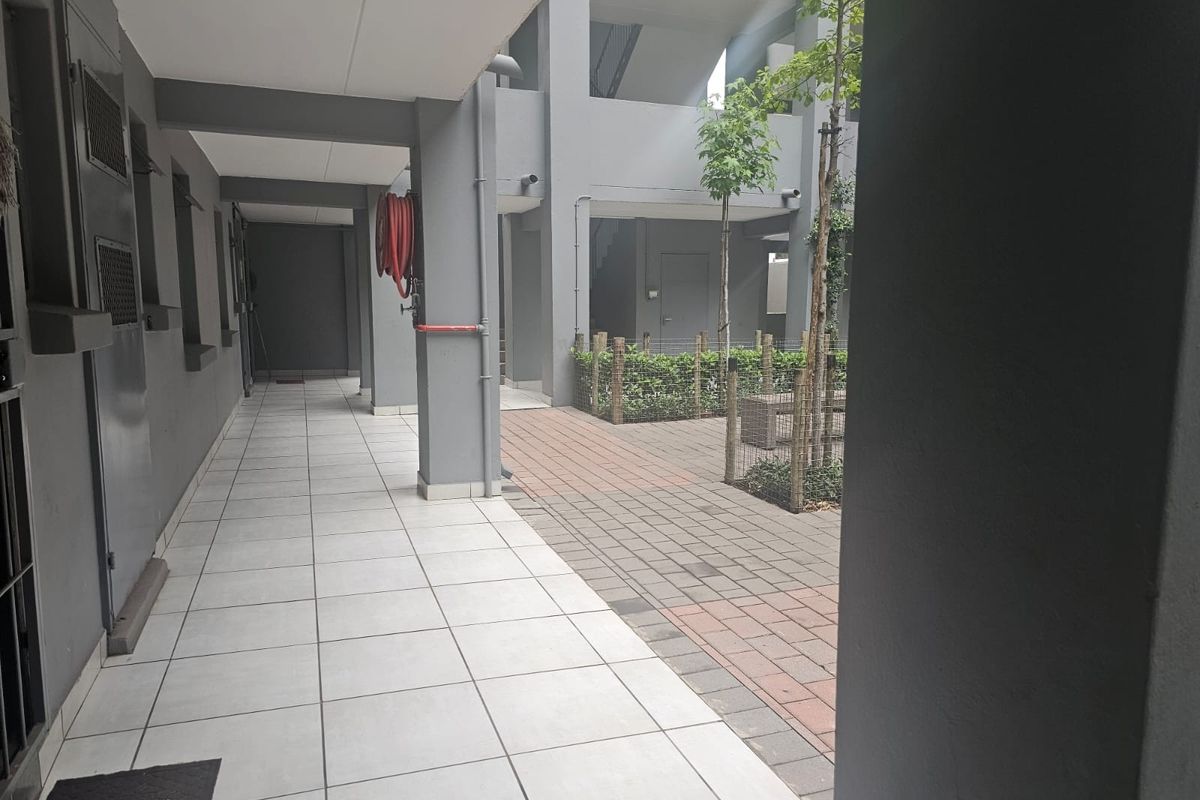 A covered tiled walkway leading to a paved courtyard area within a modern townhouse complex, featuring concrete structural pillars and a fire hose reel.