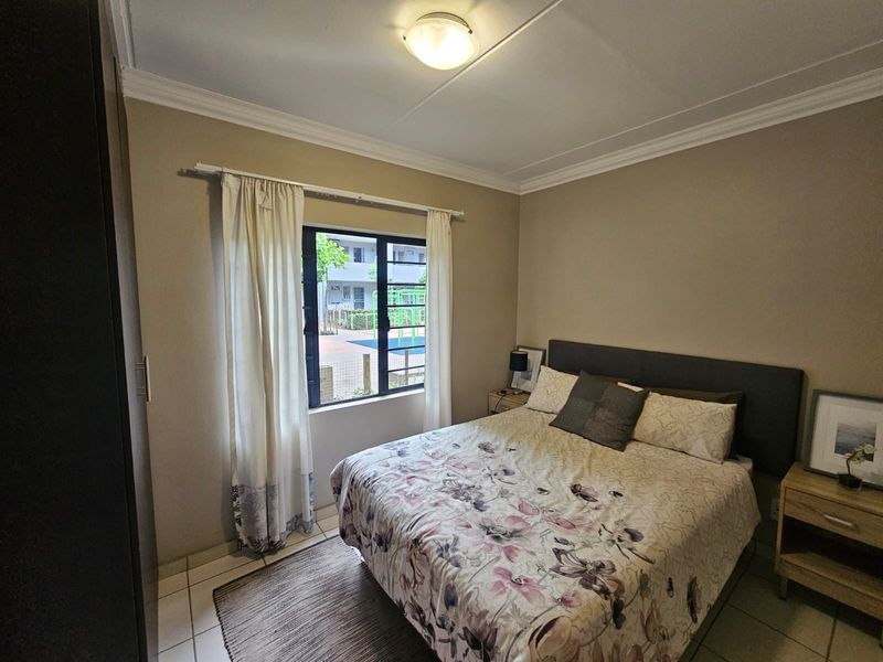 A functional bedroom featuring tiled flooring, a built-in wardrobe, and a window providing natural light and views of the complex grounds.