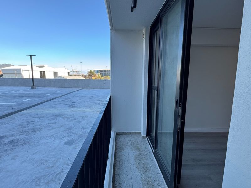 A compact balcony featuring tiled flooring and a metal railing, accessible via a sliding glass door, overlooking an adjacent concrete parking area.