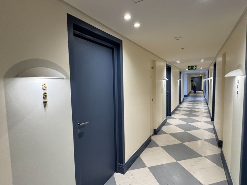 A well-maintained apartment building hallway featuring checkered flooring, recessed lighting, and uniform door frames with unit numbering.