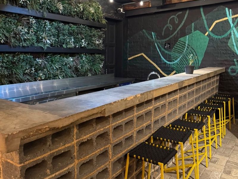 A stylish, industrial-themed bar area featuring a concrete block counter, stainless steel prep space, vertical garden wall, and exposed timber beam ceiling.