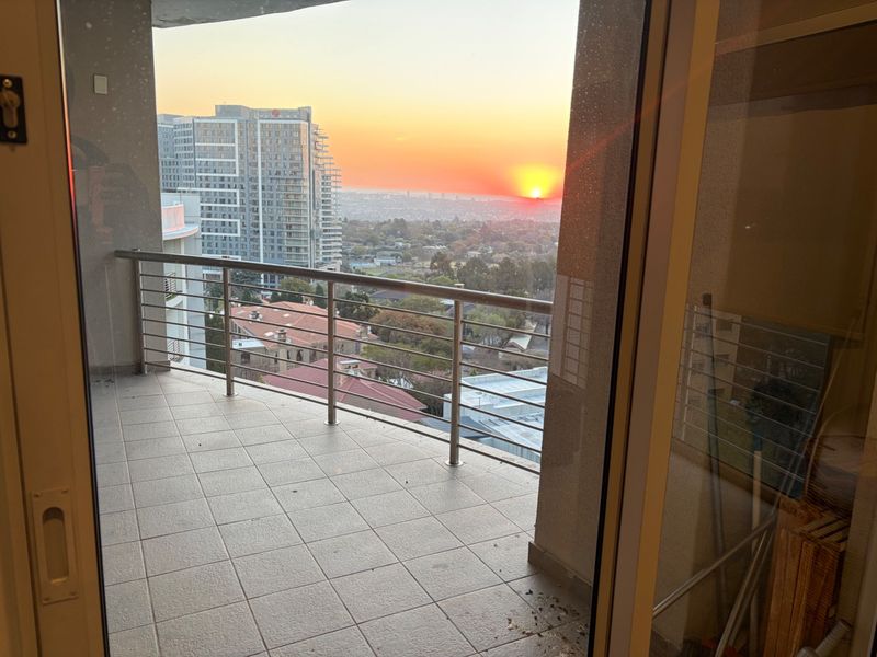A balcony with tiled flooring and a stainless steel balustrade, offering an elevated sunset view over the surrounding urban landscape.