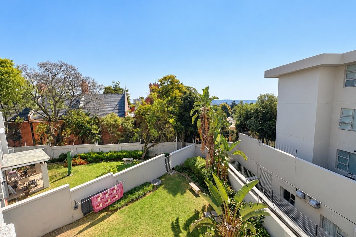An elevated view of the rear garden area featuring a well-maintained lawn, perimeter walls with security fencing, and a paved patio section.