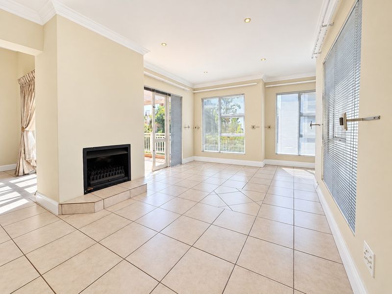Spacious, light-filled living room featuring neutral tiled flooring, a built-in fireplace, and large windows with venetian blinds, leading out to a balcony.