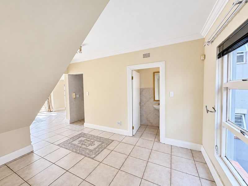 An empty living area featuring neutral tiled flooring with a decorative inlay, crown molding, and access to an adjoining bathroom space.