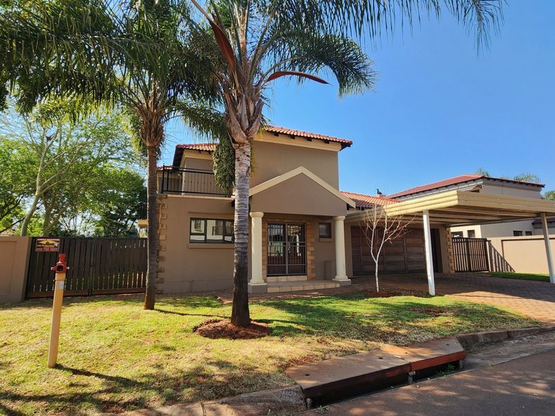Front elevation of a two-story house featuring a paved driveway, carport, balcony, and established garden with palm trees.