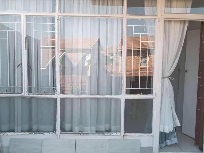 A covered balcony area featuring large glass windows with security bars, tiled flooring, and exposed brickwork, showing signs of wear on the window frames.