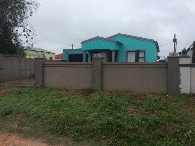 Front view of a single-story house featuring a bright teal facade, a covered entrance, and a surrounding perimeter wall with a metal gate.