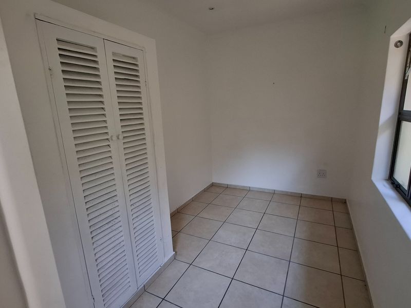 An empty bedroom featuring tiled flooring, a built-in cupboard with louvered doors, and a window fitted with security bars.