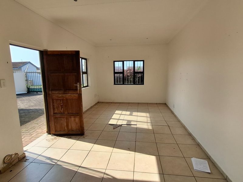 An empty, open-plan living area featuring tiled flooring, security-barred windows, and direct access to the exterior through a timber door.