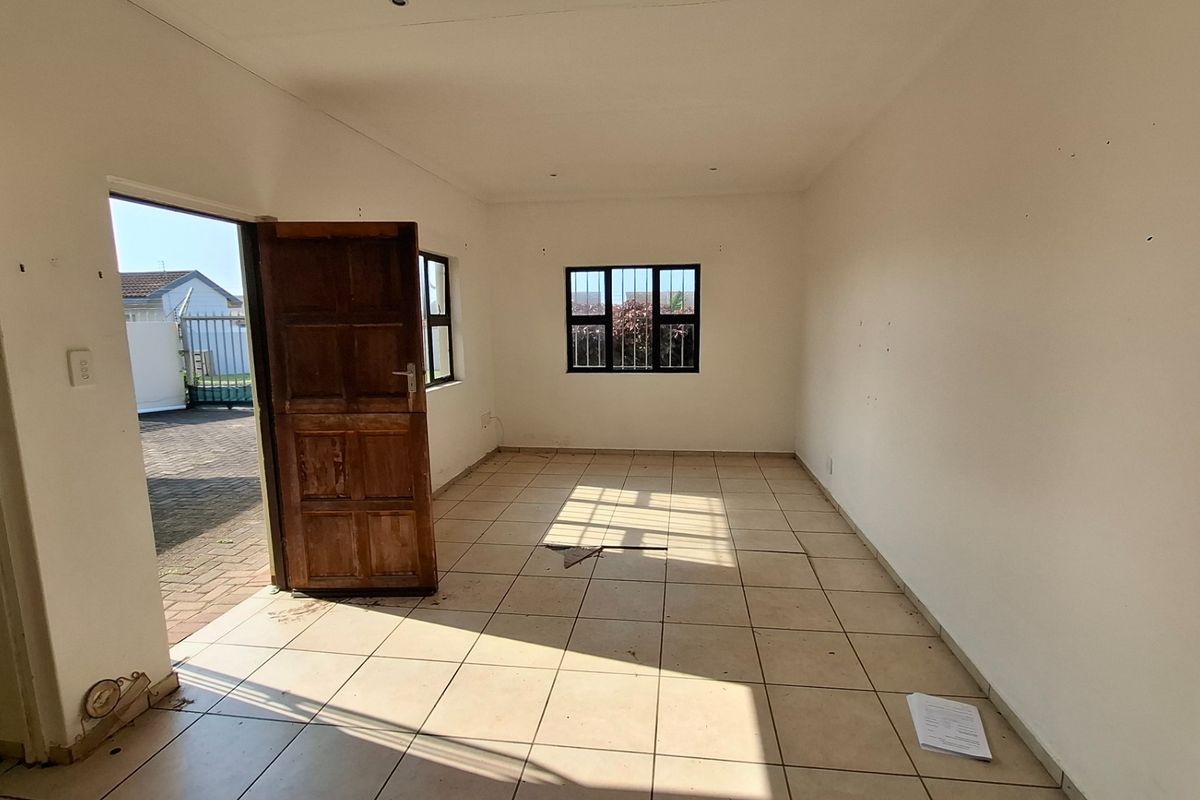 An empty, open-plan living area featuring tiled flooring, security-barred windows, and direct access to the exterior through a timber door.