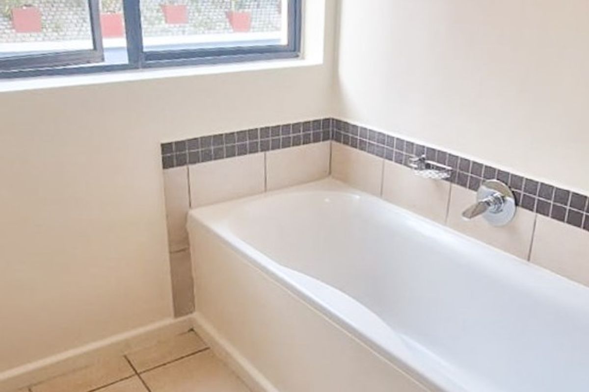 A clean, empty bathroom featuring a built-in bathtub with tiled splashback, neutral floor tiles, and a large window providing natural light.
