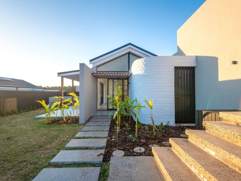 Modern rear exterior featuring a unique curved white brick wall, timber pergola, and concrete pathway leading to a contemporary home with pitched roofline.