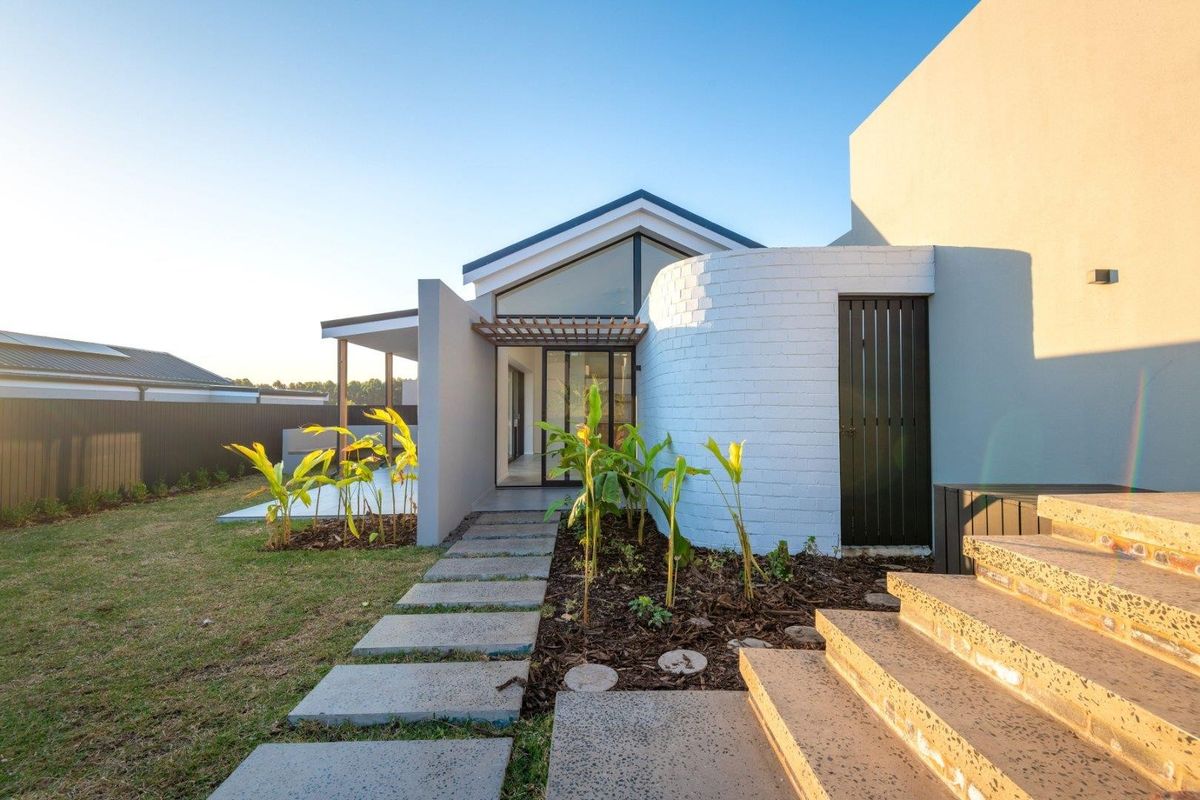 Modern rear exterior featuring a unique curved white brick wall, timber pergola, and concrete pathway leading to a contemporary home with pitched roofline.