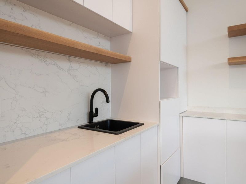 Modern scullery featuring sleek white cabinetry, stone countertops, marble-look splashback, and contemporary black fixtures with floating timber shelving.