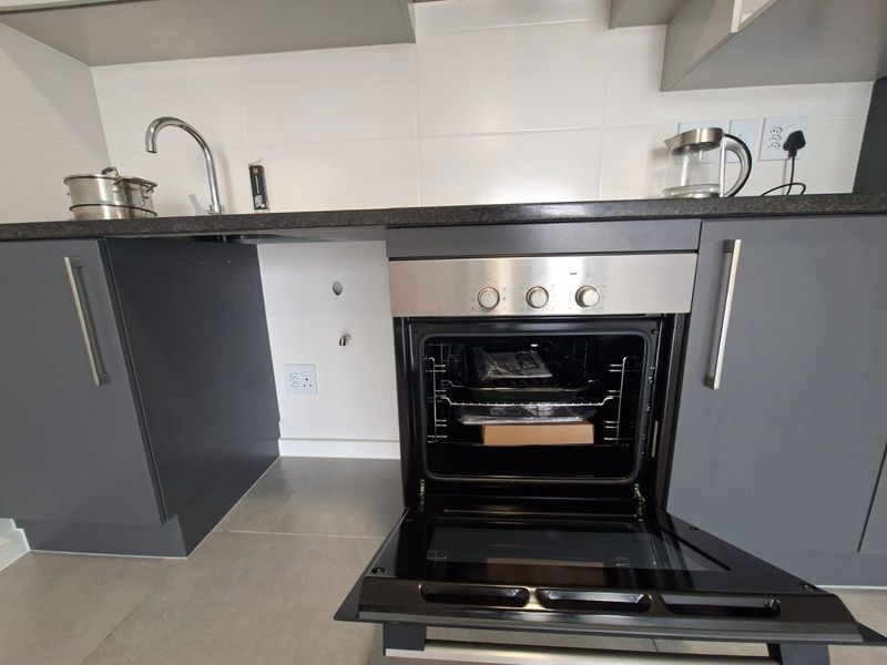 A modern kitchen featuring grey cabinetry, a built-in oven, dark countertops, and a tiled backsplash with a stainless steel sink.
