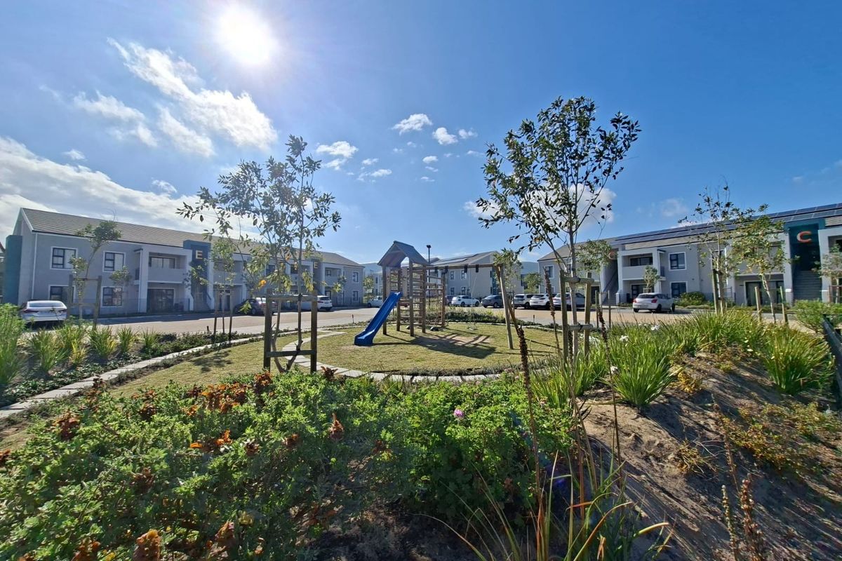 A well-maintained communal garden area featuring a children's playground, landscaped greenery, and paved parking spaces between apartment blocks.
