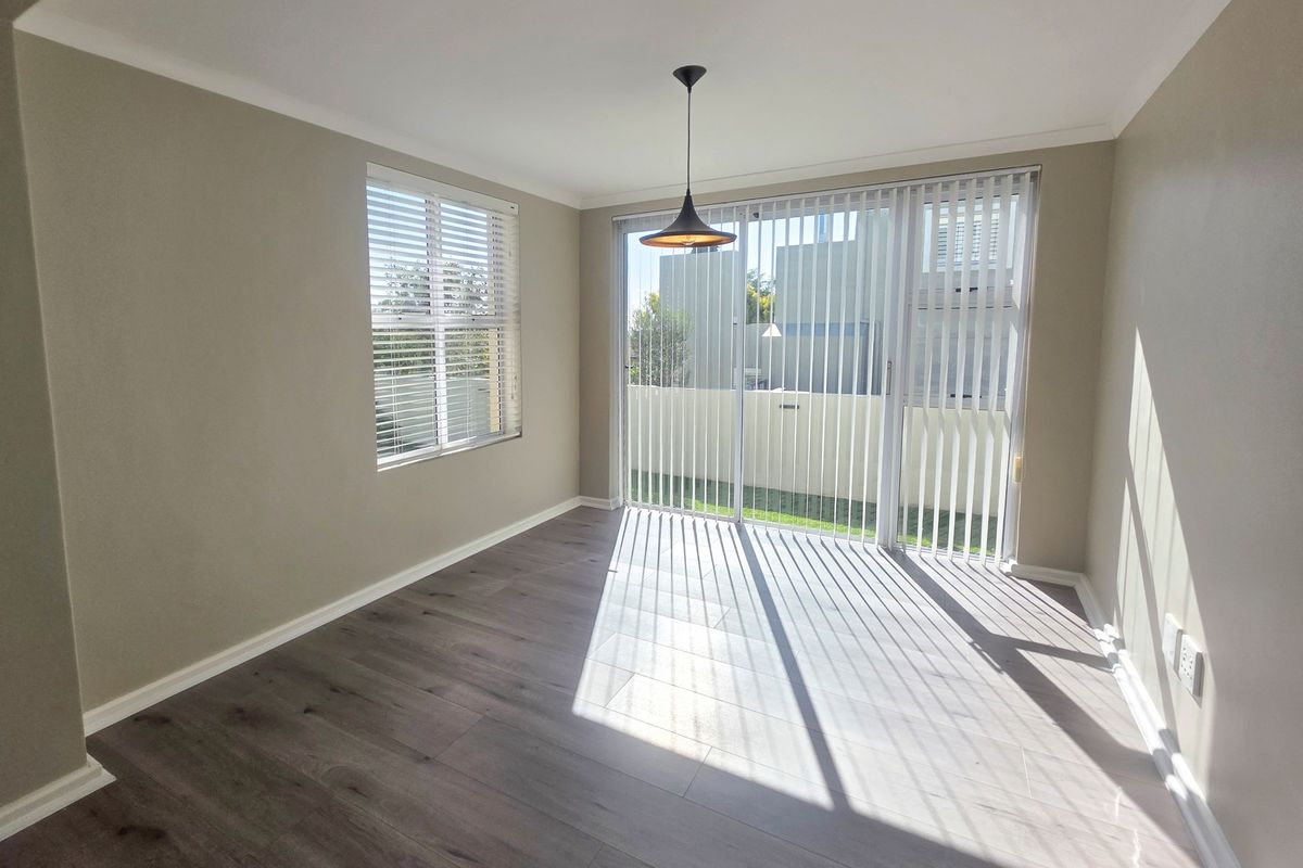 A bright, empty living room featuring modern laminate flooring, a pendant light, and large sliding glass doors with vertical blinds leading to the exterior.