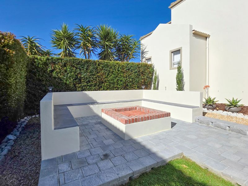A well-maintained rear garden area featuring a paved patio, built-in brick fire pit, and integrated bench seating surrounded by a manicured hedge.