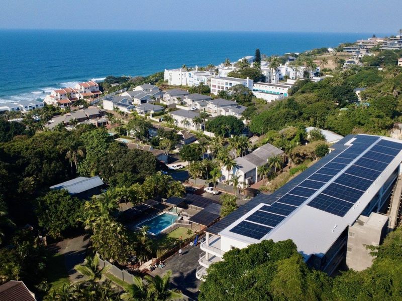 Aerial view of a coastal residential complex featuring a building with extensive rooftop solar panels, a communal swimming pool, and lush surrounding greenery.