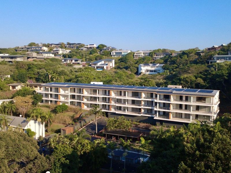 Aerial view of a modern multi-story apartment complex nestled in a lush, green hillside setting with solar panels installed on the roof.