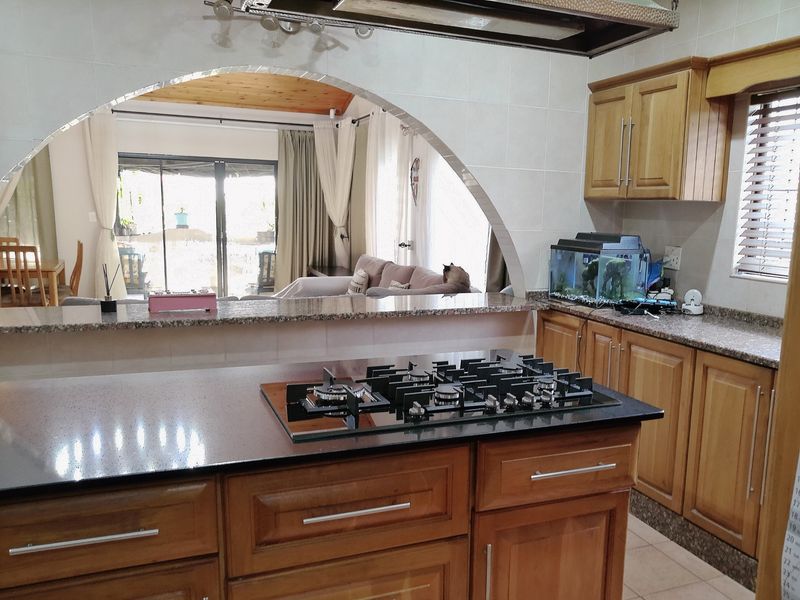 Open plan kitchen featuring wooden cabinetry, granite countertops, and a central island with a gas hob, overlooking the adjacent living area through an archway.